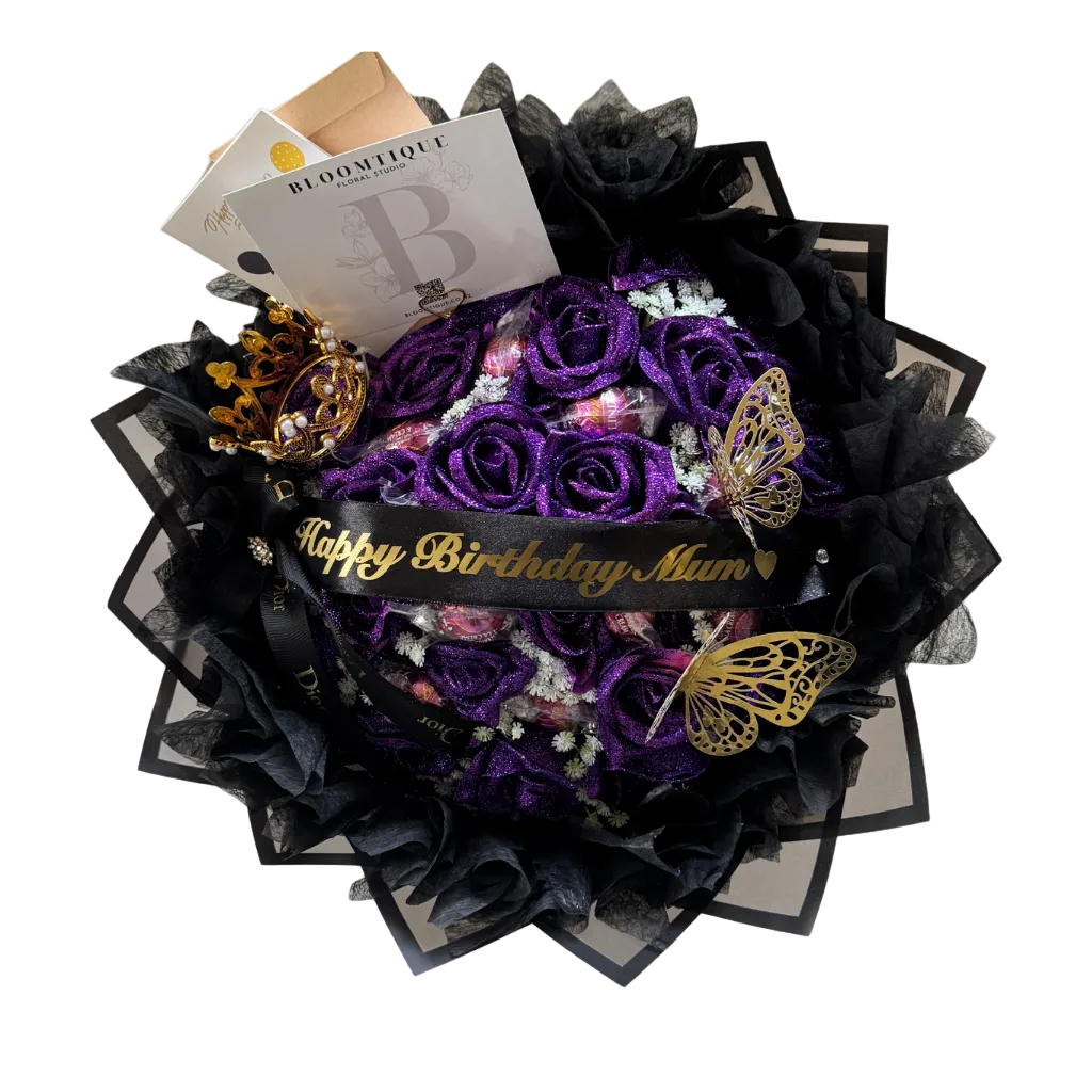 A spherical bouquet of deep purple roses wrapped in black tulle and black paper, with a black ribbon reading 'Happy Birthday Mum' in gold script. Decorative gold butterfly ornaments, a small gold crown, and a Boumontfør card are arranged around the bouquet on a white background.