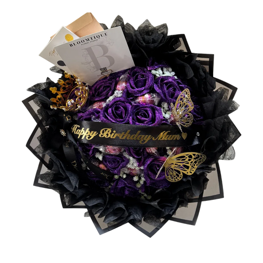 A spherical bouquet of deep purple roses wrapped in black tulle and black paper, with a black ribbon reading 'Happy Birthday Mum' in gold script. Decorative gold butterfly ornaments, a small gold crown, and a Boumontfør card are arranged around the bouquet on a white background.
